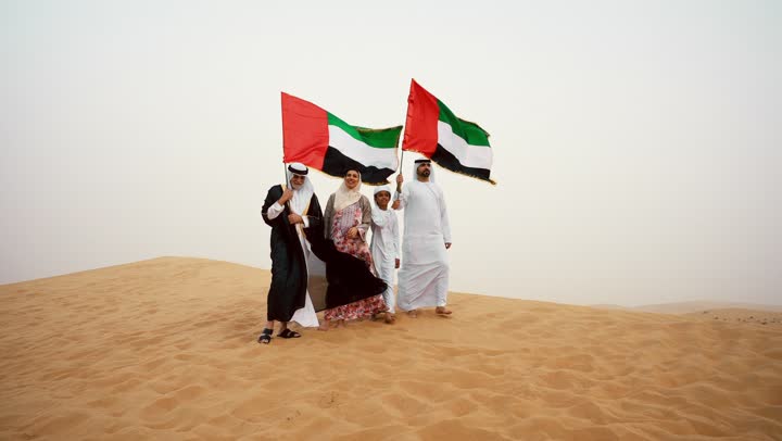 Heritage atmosphere reflecting authenticity and national belonging, the concept of pride and national unity, celebrating national identity and belonging, an Arab Gulf Emirati family wearing traditional attire standing in the desert and raising the flag of the United Arab Emirates, looking at something.