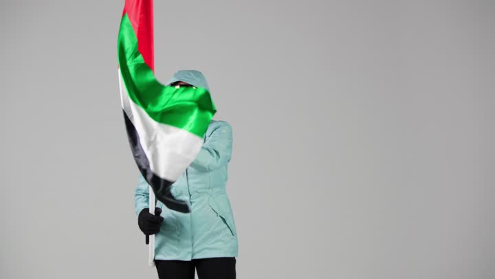 An Emirati woman wearing ski clothes holds the Emirati flag, enjoying the cold weather, the green flag fluttering high in the sky, feeling warm.