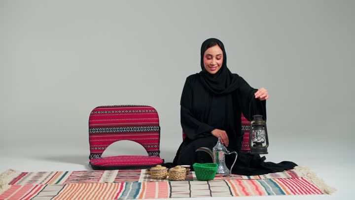 رفع المصباح المضئ للاعلى اشارة الى اقتراب الشهر الفضيل , امرأة عربية خليجية سعودية محجبة ترتدي العباءة السوداء تجلس على السجادة الملونة , امرأة اماراتية , ايماءات السعادة و الفرحة