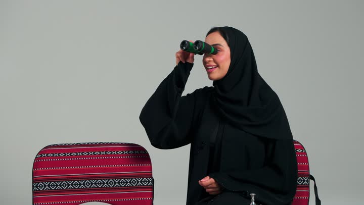 امرأة عربية خليجية سعودية محجبة ترتدي العباءة السوداء تمسك المنظار المقرب باليدين , النظر الي شئ بعيد متحرك , الاشارة باصبع السبابة بايماءات المفاجأة و البهجة الشديدة , امراة اماراتية