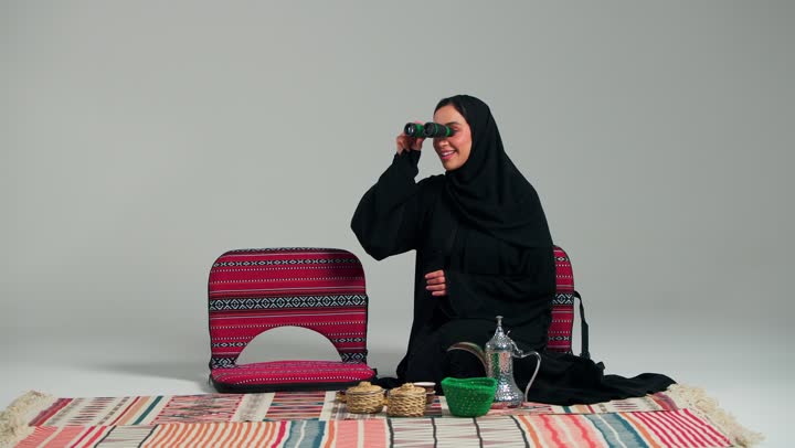 ايماءات السعادة و الفرحة الشديدة اثناء النظر في المنظار المقرب للاشياء البعيدة , امراة عربية خليجية سعودية محجبة ترتدي العباءة السوداء تشير باصبع السبابة الي شئ ما بعيد , اجواء شتوية دافئة و لحظات مميزة سعيدة , امراة اماراتية