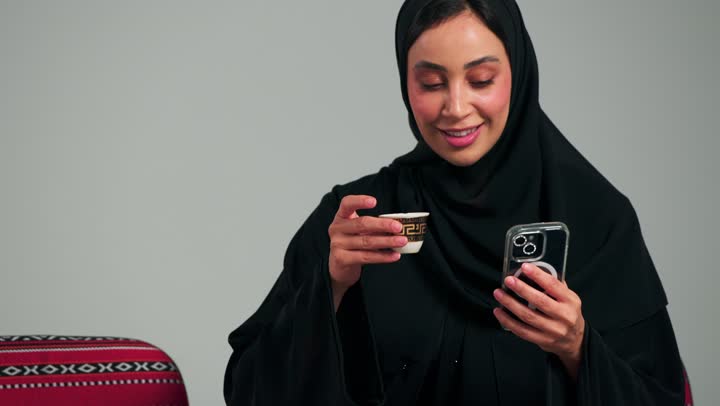 احتساء القهوة العربية بسعادة و الاستمتاع بالطعم الشهي و النكهة القوية , امرأة عربية خليجية سعودية محجبة ترتدي عباءة سوداء تتصفح العاتف المحمول , ايماءات السعادة و الابتسامة , تصوير مقرب , امرأة اماراتية