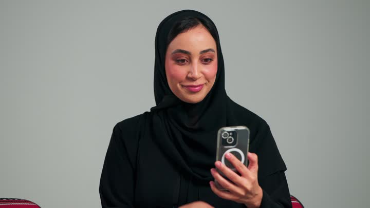 السؤال عن الصحة و الاحوال اثناء مكالمة الفيديو المباشرة , امرأة عربية خلجية سعودية محجبة ترتدي عباءة سوداءتقوم بالتلويح بكف اليد اشارة الترحيب و السلام , الجلوس على الارض بسعادة و ابتسامة , امراة اماراتية