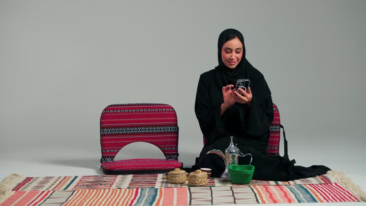 الجلوس على الارض اثناء التخييم , امرأة عربية خليجية سعودية محجبة ترتدي عباءة سوداء تستخدم الهاتف المحمول للقيام بالتواصل المباشر مع الاهل و الاصدقاء , التلويح بكف اليد في بداية مكالمة الفيديو , ايماءات السعادة و السرور ,امراة اماراتية