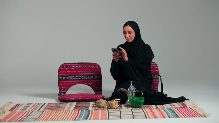 النظر الى شئ ما , امرأة عربية خليجية سعودية محجبة ترتدي عباءة سوداء تجلس على الارض , استخدام الهاتف المحمول للتسلية و قضاء الوقت الممتع , امرأة اماراتية