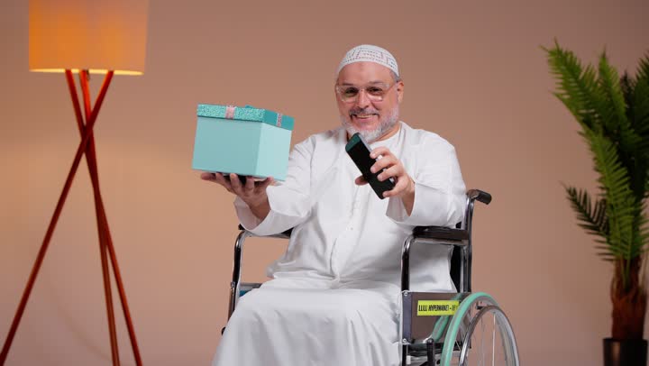 An elderly Arab Gulf Saudi man wearing a white thobe and a white cap is holding a blue gift box, showing a blank green mobile phone screen to the camera, sitting in a wheelchair, with expressions of surprise and happiness.