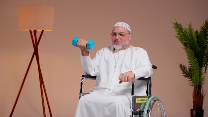 النظر للكاميرا بسعادة اثناء ممارسة التمارين الرياضية , رجل عربي خليجي سعودي مسن يرتدي الثوب الابيض و الطاقية البيضاء يحمل الاوزان الحديدية بسعادة , الحصول على جسد قوي , الجلوس على الكرسي المتحرك , ايماءات الفرحة و السرور