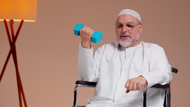 تقوية عضلات الذراع للحصول على جسد قوي , رجل عربي خليجي سعودي مسن يرتدي الثوب الابيض و الطاقية البيضاء يحمل الاوزان الحديدية الزرقاء , الجلوس على الكرسي المتحرك بسعادة , النظر للكاميرا بابتسامة