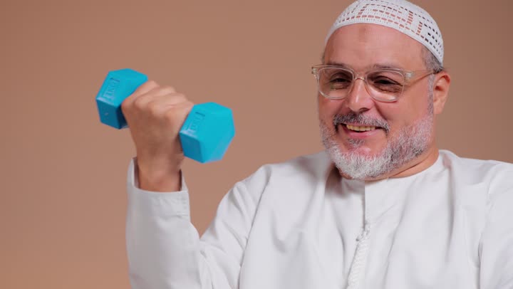 ثني الذراع مراراً و تكراراً للحصول على جسد قوي , رجل عربي خليجي سعودي مسن يرتدي الثوب الابيض و الطاقية البيضاء يمارس التمارين الرياضية بسعادة , تصوير مقرب , الابتسامة اثناء ممارسة الرياضة