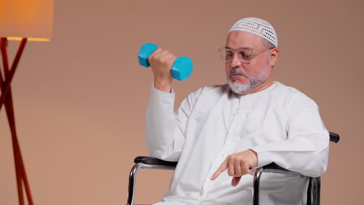 ممارسة الرياضة بسعادة , رجل عربي خليجي سعودي مسن يرتدي الثوب الابيض و الطاقية البيضاء يحمل الاوزان الثقيلة , الاشارة باصبع السبابة لعضلات الذراع , ايماءات الفرحة و السعادة , الجلوس على الكرسي المتحرك