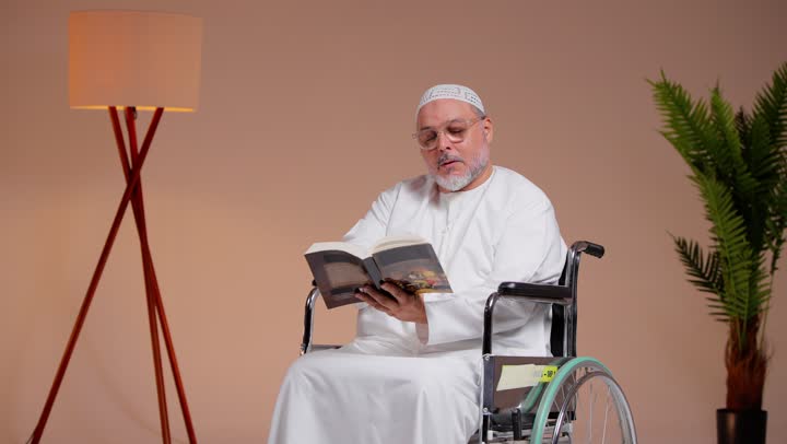 Increasing knowledge and information through useful reading, an elderly Arab Gulf Saudi man wearing a white cap and a white thobe holding an interesting book, sitting in a wheelchair, gestures of joy and happiness.