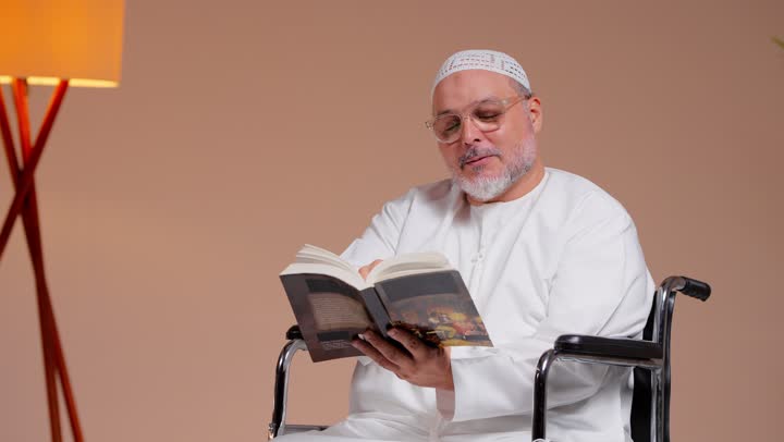 القراءة الممتعة المفيدة للعقل , رجل عربي خليجي سعودي مسن يرتدي الثوب الابيض و الطاقية البيضاء يحمل كتاب , الجلوس على الكرسي المتحرك , ايماءات السعادة و الفرحة