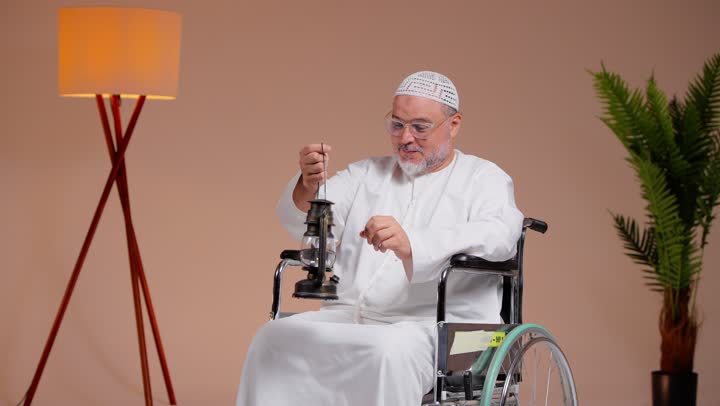 اضاءة فانوس رمضان اشارة الى اقتراب الشهر الفضيل , رجل عربي خليجي سعودي مسن يرتدي الطاقية البيضاء و الثوب الابيض يجلس على الكرسي المتحرك , ايماءات السعادة و الابتسامة