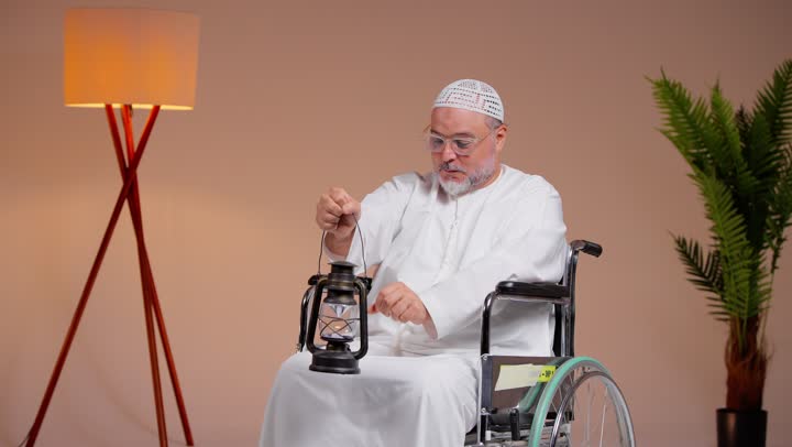 رجل عربي خليجي سعودي مسن يرتدي الثوب الابيض و الطاقية البيضاء يحمل مصباح مضئ بسعادة و سرور , الجلوس على الكرسي المتحرك , ايماءات الفرحة و البهجة لاقتراب شهر رمضان المبارك ,