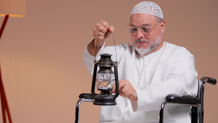 رفع المصباح المضئ بسعادة احتفالاً بقدوم الشهر الفضيل , رجل عربي خليجي سعودي يرتدي الثوب الابيض و الطاقية البيضاء يجلس على الكرسي المتحرك , ايماءات السعادة و الابتسامة امام الكاميرا , تصوير مقرب