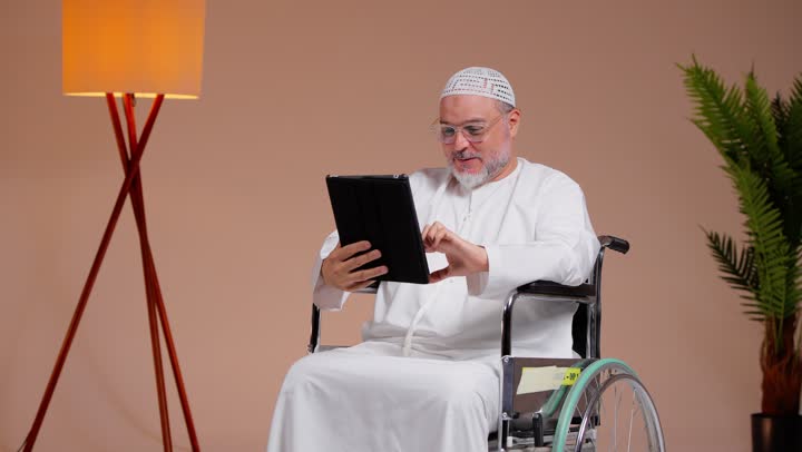 مشاهدة صور العائلة و الاحفاد الصغار  بفرحة عارمة , رجل عربي خليجي سعودي يرتدي الثوب الابيض و الطاقية البيضاء يتصفح الجهاز اللوحي  , الجلوس على الكرسي المتحرك , ايماءات البهجة و السعادةلوحي