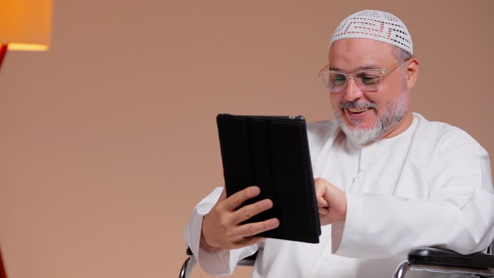 الاطمئنان على اخبار العائلة من اي مكان بسهولة باستخدام التطبيقات المتعددة لمحادثات الفيديو المباشرة , رجل عربي خليجي سعودي يرتدة ثوب ابيض و طاقية بيضاء يجري مكالمة عائلية , ايماءات السعادة و الابتسامة للجهاز اللوحي , الجلوس على الكرسي المتحرك
