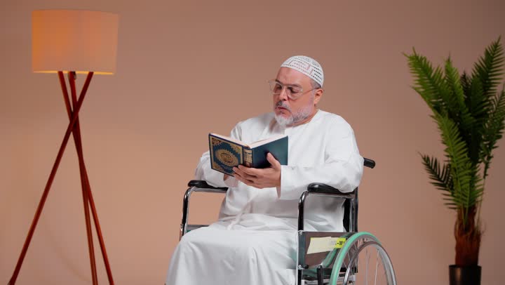 قراءة ايات الرحمن و الشعور بالسكينة و راحة القلب , رجل عربي خليجي سعودي يرتدي الثوب الابيضو الطاقية البيضاء يمسك المصحف الشريف , الجلوس على الكرسي المتحرك