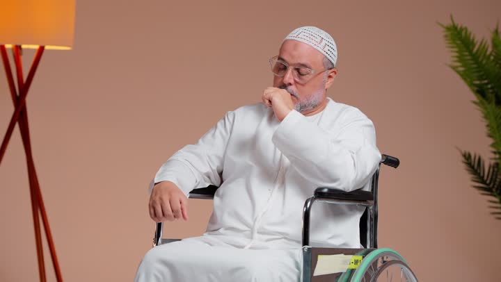 توجيه الجهاز الى الرأس , رجل عربي خليجي سعودي يرتدي الثوب الابيض و الطاقية البيضاء يضغط على الجبهة بسبب الشعور بالألم , طبيب يرتدي المعطف الطبي و القفازات الزرقاء يسجل القراءة للاطمئنان على المريض , الجلوس على الكرسي المتحرك