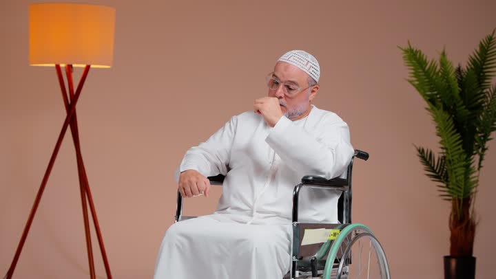 قياس درجة حرارة الجسم و الاطمئنان على الصحة العامة للمريض , رجل عربي خليجي سعودي مسن يرتدي الثوب الابيض و الطاقية البيضاءيجلس على الكرسي المتحرك , الضغط على جبهة الراأس اشارة الي الشعور بالالم الشديد