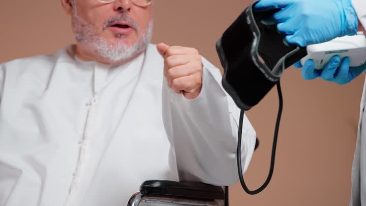 الاطمئنان على الصحة , رجل عربي خليجي سعودي مسن يرتدي طاقية بيضاء و ثوب ابيض يجلس على الكرسي المتحرك , الطبيب يستخدم الجهاز الطبي و يقيس ضغط الدم , ربط الجهاز على الذراع , رفع اصبع الابهام للاعلى علامة على القراءة الجيدة , تصوير مقرب
