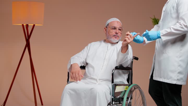 قياس نسبة الاكسجين في الدم , رجل عربي خليجي سعودي مسن يرتدي ثوب ابيض و طاقية بيضاء جلس على الكرسي المتحرك , طبيب يرتدي معطف طبي ابيض و قفازات جلدية زرقاء يرفع اصبع الابهام علامة على الاخبار الجيدة بعد الاطمئنان على الصحة , ايماءات الراحة و الاطمئنان