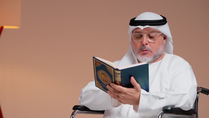 الجلوس على الكرسي المتحرك و قراءة ماتيسر من ايات الذكر الحكيم , رجل عربي خليجي سعودي يرتدي ثوب ابيض و غترة بيضاء يحمل المصحف الشريف ,التقرب الى الله , ايماءات السكينة والسلام