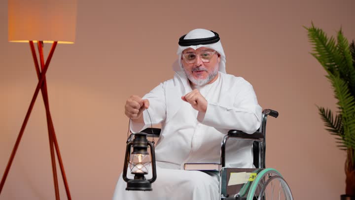 رفع اصبع الابهام لاعلى مع الابتسامة اشارة الى الفرحة و البهجة , رجل عربي خليجي سعودي مسن يرتدي غترة بيضاء و ثوب ابيض يحمل مصباح مضئ , الاحتفال بالشهر المبارك , الجلوس على الكرسي المتحرك , ايماءات السعادة