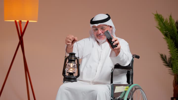 عرض شاشة هاتف محمول خضراء فارغة للكاميرا , رجل عربي خليجي سعودي مسن يرتدي ثوب ابيض و غترة بيضاء يجلس على الكرسي المتحرك , رفع الفانوس المضئ احتفالاً بالشهر الكريم , ايماءات السعادة و الفرحة