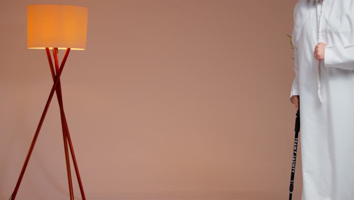 An elderly Arab Gulf Saudi man wearing a white thobe and a white ghutrah is holding onto a wooden cane while moving, close-up shot, trying to maintain balance while walking.