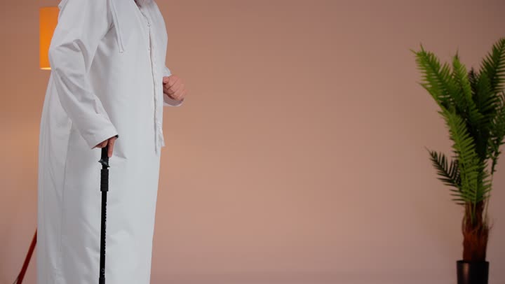Multiple tools to assist the elderly in movement, an elderly Arab Gulf Saudi man wearing a white thobe and white ghutrah leaning on a wooden cane, trying to stay balanced while walking.