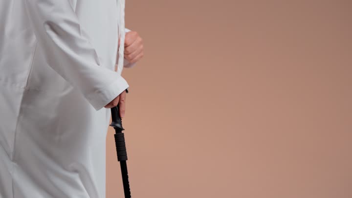 Slow movement while walking, an elderly Arab Gulf Saudi man wearing a white thobe using a wooden cane to walk, trying to regain balance, close-up shot.