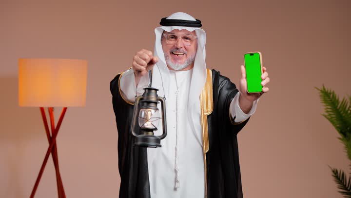 الوقوف امام الكاميرا و عرض شاشة هاتف محمول خضراء فارغة , مسن عربي خليجي سعودي يرتدي الثوب الابيض التقليدي في المملكة العربية السعودية يرفع المصباح المضئ اشارة الى قدوم شهر رمضان المبارك , ايماءات الفرحة و البهجة , غترة بيضاء ناصعة و مشلح اسود انيق ,