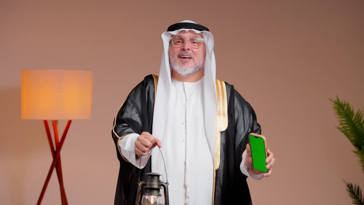 الاحتفال بقدوم شهر رمضان الفضيل , رجل عربي خليجي سعودي مسن يرتدي الثوب الابيض و البشت الاسود يحمل الفانوس المضئ , عرض شاشة هاتف محمول خضراء فارغة امام الكاميرا , ايماءات السعادة و الفرحة ,