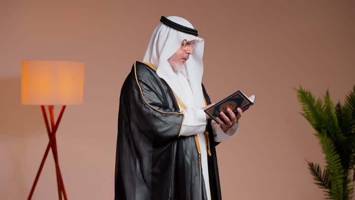 Moments of worship and drawing closer to God, an elderly Arab Gulf Saudi man wearing a white thobe and a black cloak flips through the pages of the Holy Quran, feeling the divine spirituality while reciting what is easy from the verses of the Holy Reminder.