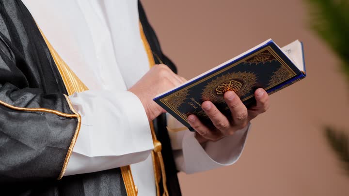 Recitation of what is easy from the verses of the Holy Quran, an elderly Arab Gulf Saudi man wearing a white ghutrah and an elegant black bisht, finishing one page and reading the other is a sign of immersion and engagement in reading.