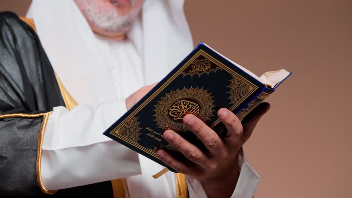 A comforting reading for the heart, an elderly Arab Gulf Omani man wearing traditional attire in the Kingdom of Saudi Arabia reads the Holy Quran, feeling tranquility, touching the page and tracing the words with his index finger.