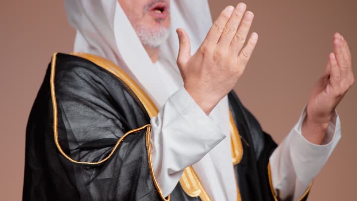 Worship and drawing closer to God through prayer and hope, an elderly Saudi Arabian Gulf man wearing the traditional white attire of the kingdom and a black cloak, raising his hands to the sky, close-up shot.
