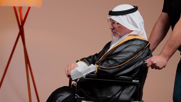 A Saudi Arabian Gulf man in casual attire helps the elderly. An elderly man sitting in a wheelchair wears a white thobe and a black cloak. There are gestures of gratitude and happiness, providing assistance and care.
