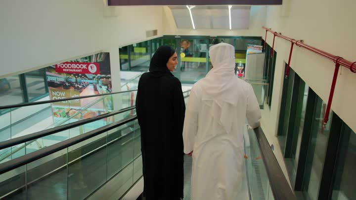 The concept of shopping and entertainment in modern centers, a rear view of an Emirati Arab couple wearing traditional attire moving on the escalator inside the mall, exchanging conversation while strolling, enjoying their time inside the shopping complexes.