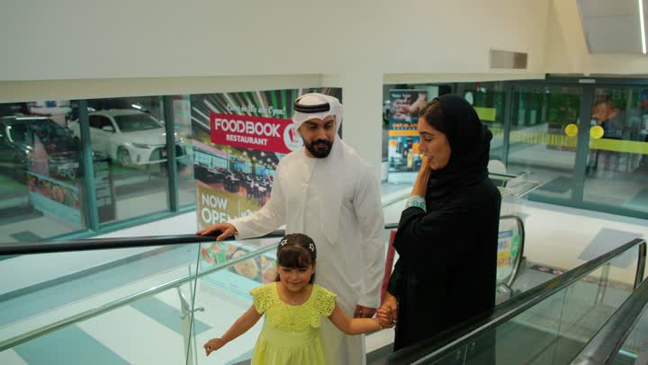 تبادل الحديث أثناء التجول، عائلة عربية خليجية إماراتية ترتدي الزي التقليدي تصعد على السلم المتحرك داخل مركز تجاري، قضاء وقت عائلي أثناء التسوق، مفهوم الترابط الاسري، ايماءات السعادة و السرور