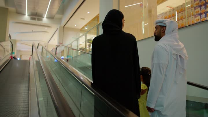 عائلة عربية خليجية إماراتية ترتدي الزي التقليدي تصعد على السلم المتحرك داخل مركز تجاري، قضاء وقت عائلي أثناء التسوق، مفهوم الترابط الاسري، ايماءات السعادة و السرور، تبادل الحديث أثناء التجول