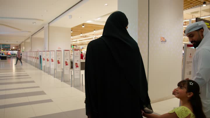 مفهوم التسوق الأسري، قضاء وقت عائلي والتنزه داخل المول، تصوير من الخلف لعائلة عربية خليجية إماراتية ترتدي الزي التقليدي داخل مركز تجاري، أب يرتدي الكندورة والغترة يسير برفقة زوجته وابنتهما، تواصل وترابط عائلي، الاستعداد لشراء الاحتياجات المنزلية