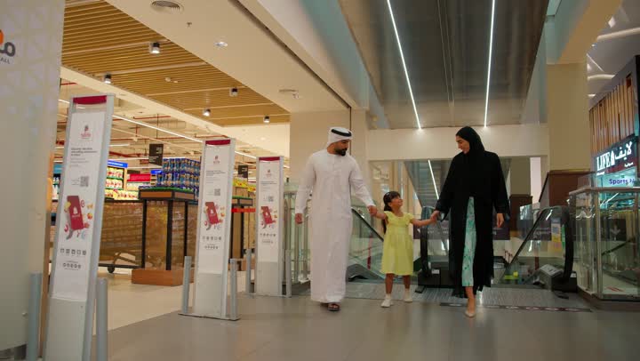 Spending family time and strolling inside the mall, an Arab Gulf Emirati family wearing traditional attire inside a shopping center, a father wearing a kandura and ghutrah walking with his wife and their daughter, the concept of family shopping, communication and family bonding.