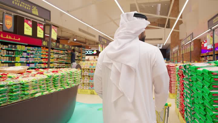 اختيار المواد الغذائية بعناية قبل الشراء، التسوق اليومي وشراء الاحتياجات المنزلية، مفهوم التسوق الذكي والتوفير، تصوير من الخلف لرجل عربي خليجي إماراتي يرتدي الكندورة والغترة يجر العربة يقوم بتفقد أكياس الأرز داخل السوبر ماركت أثناء التسوق