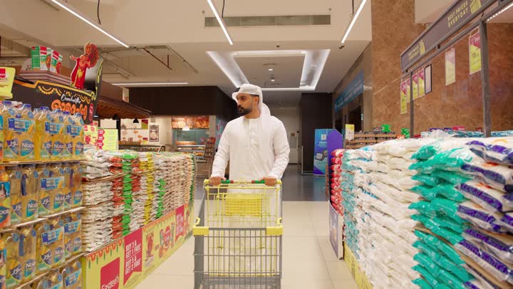 التسوق اليومي وشراء الاحتياجات المنزلية، مفهوم التسوق الذكي والتوفير، رجل عربي خليجي إماراتي يرتدي الكندورة والغترة يجر العربة يقوم بتفقد أكياس الأرز داخل السوبر ماركت أثناء التسوق، اختيار المواد الغذائية بعناية قبل الشراء