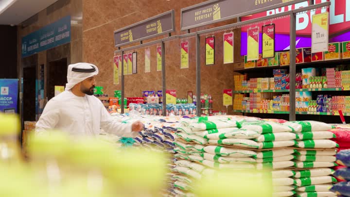 مفهوم التسوق الذكي والتوفير، رجل عربي خليجي إماراتي يرتدي الكندورة والغترة يجر العربة يقوم بتفقد أكياس الأرز داخل السوبر ماركت أثناء التسوق، اختيار المواد الغذائية بعناية قبل الشراء، التسوق اليومي وشراء الاحتياجات المنزلية