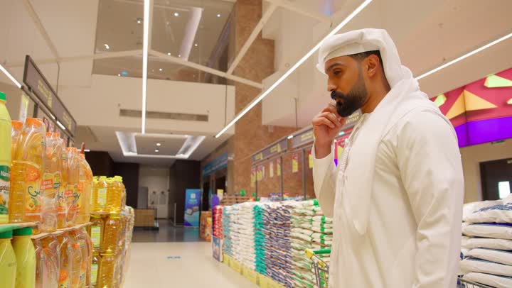 Choosing oils and food supplies, an Arab Gulf Emirati man wearing a kandura and ghutrah stands inside the supermarket carefully inspecting food products before making a purchase, smart shopping and making a buying decision, the concept of daily shopping, buying household necessities.