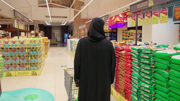 شراء الأرز والبقالة، تصوير من الخلف لامرأة عربية خليجية إماراتية محجبة ترتدي العباءة تقوم بدفع عربة التسوق داخل السوبر ماركت وشراء المواد الغذائية والتموينية، مفهوم التسوق اليومي، اختيار الاحتياجات المنزلية