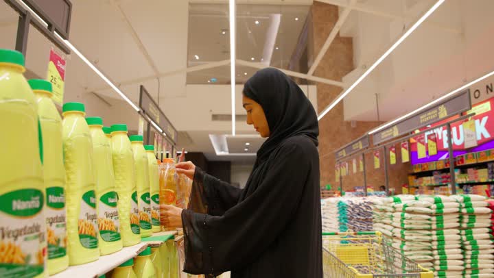 Making a purchase decision, the concept of smart shopping, an Arab Gulf Emirati woman wearing a hijab and an abaya stands inside the supermarket, thinking and carefully selecting food products from the shelves, buying daily necessities.